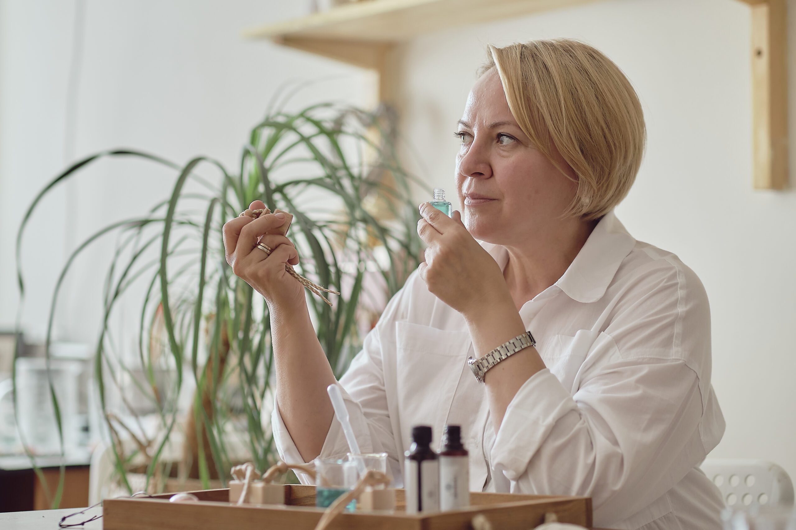 Mulher com roupas brancas cheira um pequeno frasco de líquido azul enquanto segura um ingrediente natural com a outra mão, em um ambiente que sugere a avaliação e preparação de terapias naturais ou produtos de bem-estar.