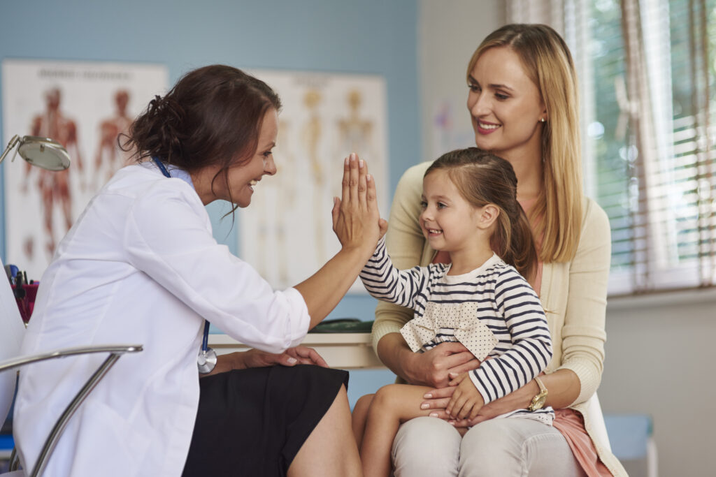Neuropediatra em uma consulta acolhedora dá um "high-five" para uma menina sorridente que está no colo da mãe, demonstrando um ambiente de confiança para a avaliação neurológica infantil.