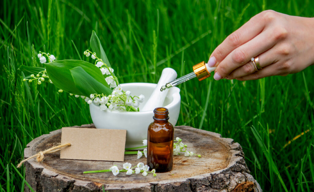 Mão segurando um conta-gotas com líquido, prestes a pingar em um frasco âmbar, ao lado de um gral com lírios do vale e folhas verdes, simbolizando a extração e o preparo cuidadoso de medicamentos homeopáticos naturais.