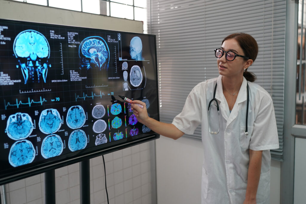 Médica neurologista analisa múltiplos exames de imagem do cérebro, como ressonância magnética, em uma tela grande para realizar um diagnóstico neurológico preciso.
