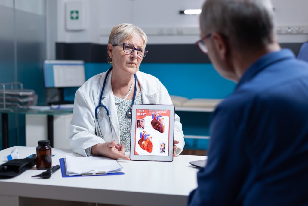 Cardiologista experiente explica a um paciente sobre os riscos de um infarto usando um tablet com imagens do coração, ilustrando a importância da consulta médica.